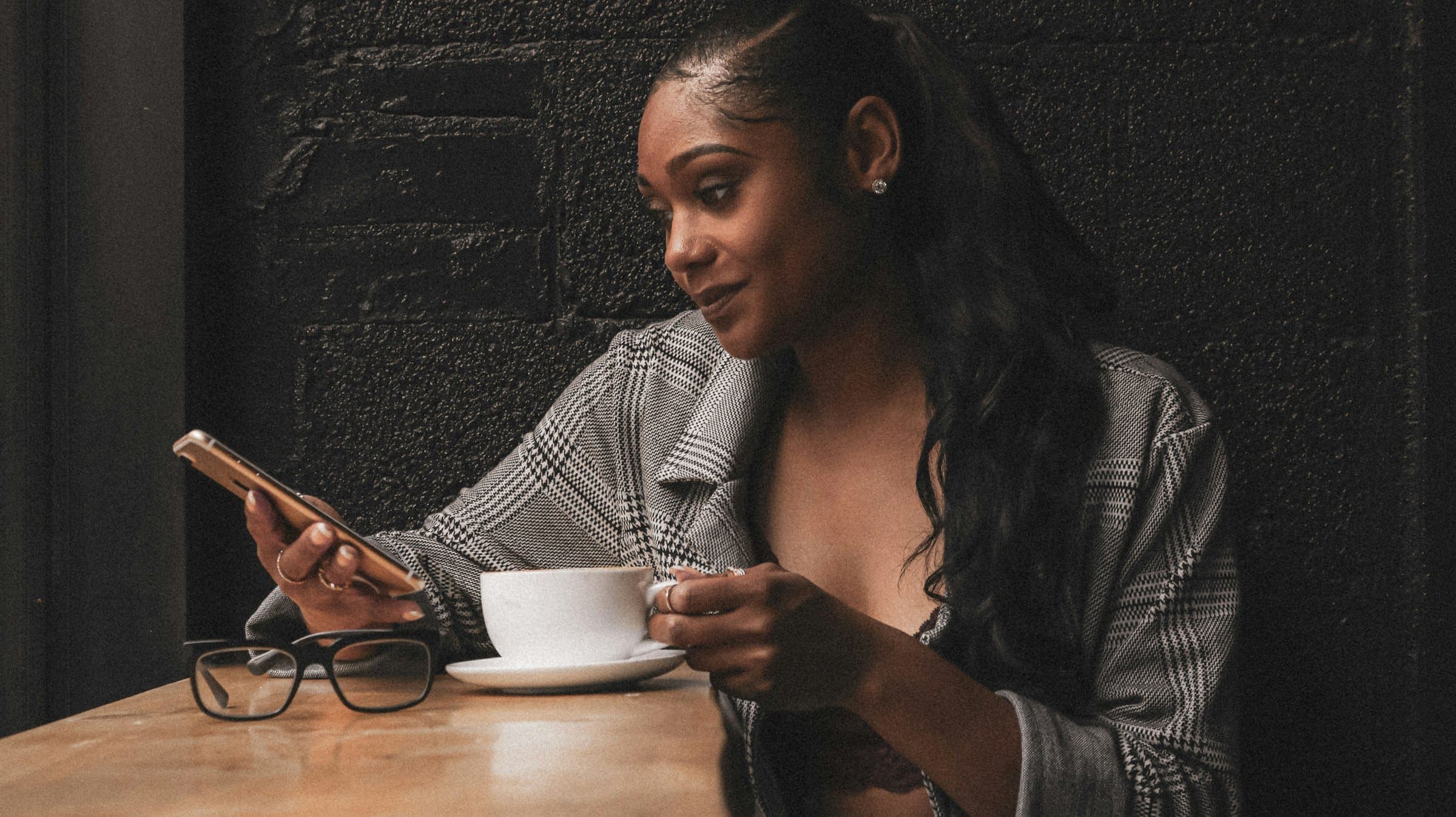 Woman sits at a café table smiling at her phone beside a cup of coffee
