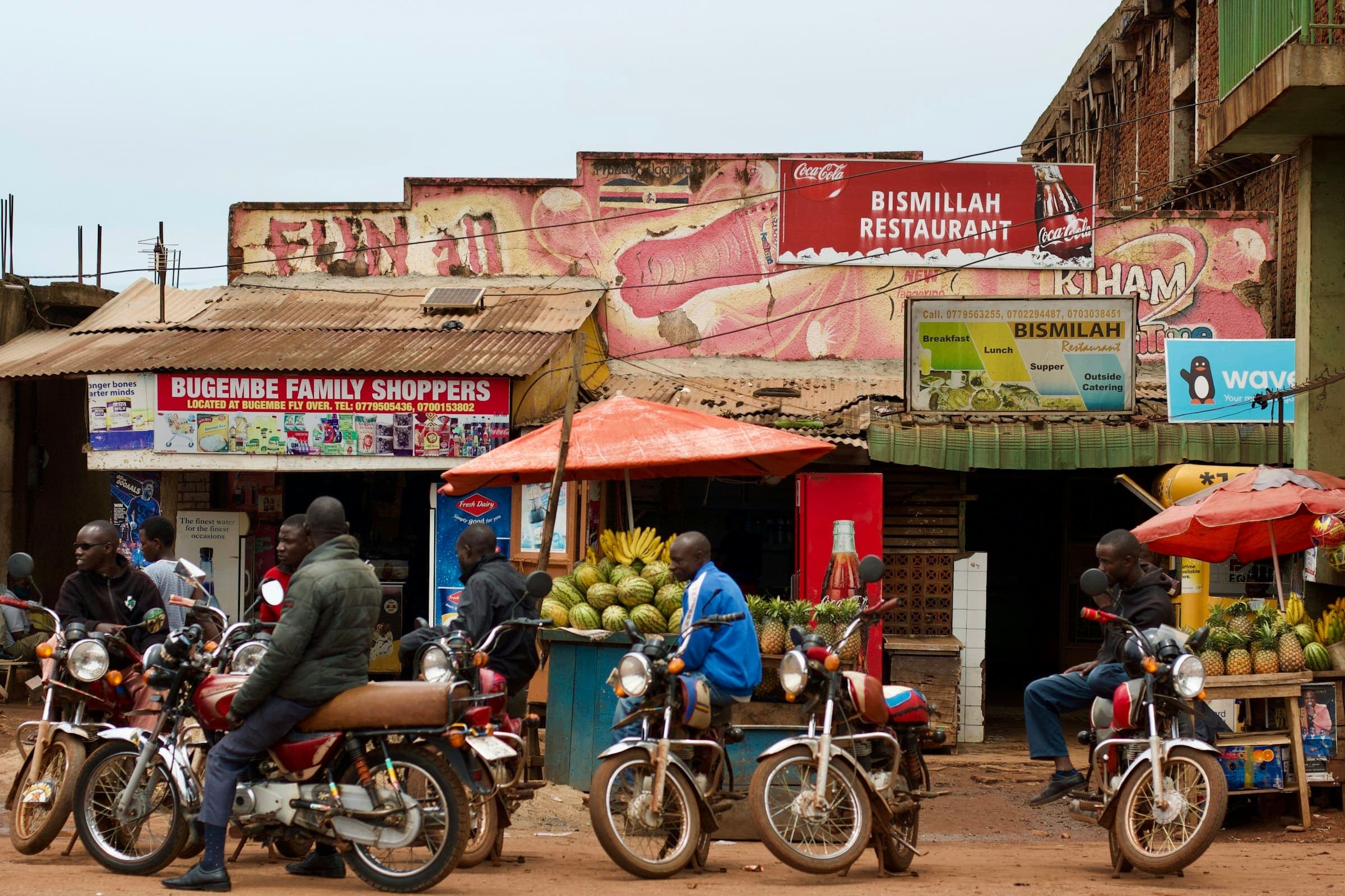 Motorcycle taxi riders wait on a busy street lined with small shops